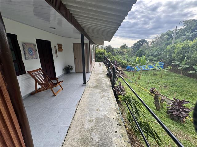 eco-hotel-gaia-2 - Alquiler de Fincas en el Quindio, Hoteles en el Eje Cafetero Concrete balcony along a white house with a wooden chair, tiled floor, and railing overlooking a tropical garden.