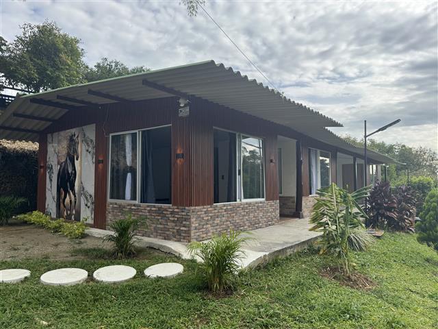 eco-hotel-gaia-3 - Alquiler de Fincas en el Quindio, Hoteles en el Eje Cafetero Single-story house with a corrugated metal roof, wood siding, brick base, large windows, and a front yard with stepping-stone path and tropical plants.
